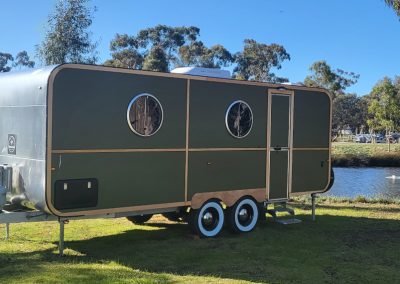 Modern green caravan parked by the lake at Stawell Hotel Motel Accommodation, featuring a stylish design with dark green and wood paneling against a clear blue sky