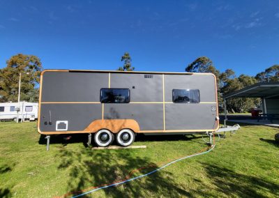 Modern grey caravan with wooden accents parked by the lake at Stawell Hotel Motel Accommodation, showcasing a sleek design against a backdrop of clear blue skies and lush greenery