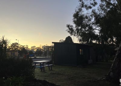 A serene sunrise over Stawell, VIC, highlighting a quiet accommodation setting with a dark building and a picnic table under soft morning light, framed by lush greenery and trees