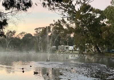 A tranquil sunrise over a misty lake in Stawell, VIC, showcasing a fountain spraying water and ducks swimming nearby, with trees framing the serene landscape and accommodation facilities visible in the background