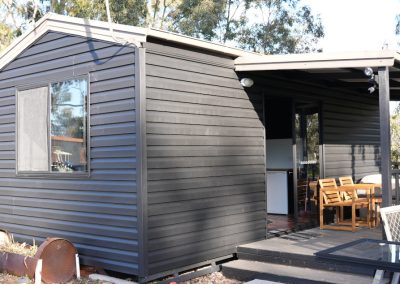 Side view of a Cabin at Stawell Hotel Motel, displaying a dark wooden exterior with a window, covered porch, and outdoor seating. The cabin is surrounded by a natural setting with greenery in the background.