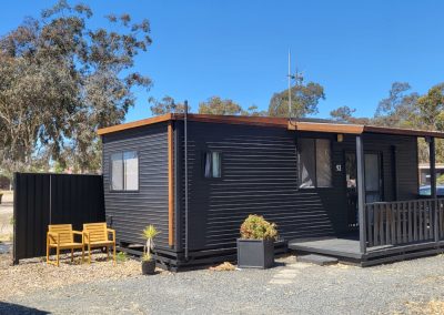 A modern black cabin at Stawell Hotel Motel Accommodation labeled with the house number "92" with horizontal siding, featuring a small covered porch with a wooden railing. Two wooden chairs and potted plants are positioned next to the entrance. The surrounding area is a campground with gravel ground and greenery, including trees in the background under a clear blue sky.