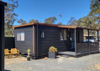 Front view of a contemporary cabin at Stawell Hotel Motel Accommodation, showcasing a dark exterior with wooden accents, a covered porch with seating, and a gravel pathway. Surrounded by lush greenery and trees, the cabin is designed for comfort and relaxation in a serene setting