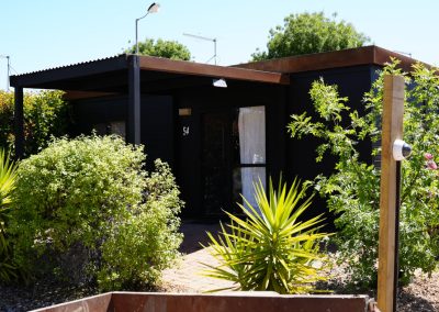 Close-up view of a Cabin at Stawell Hotel Motel Accommodation, featuring a sleek black exterior with wooden accents and a covered entrance. Lush greenery and ornamental plants surround the cabin, enhancing its inviting atmosphere under a clear blue sky