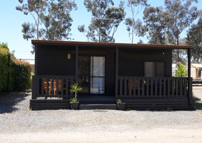 Front view of a Cabin at Stawell Hotel Motel Accommodation, showcasing a modern design with a dark exterior and wooden porch. The cabin features a spacious deck with seating, surrounded by gravel pathways and tall trees in the background, creating a serene and inviting environment