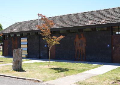 Exterior view of the main amenities building at Stawell Hotel Motel Accommodation, featuring unique metal silhouettes of two figures on the wall. The building has a textured dark facade and a corrugated roof, complemented by a wooden stump and landscaped grass area in front. Clear blue skies enhance the inviting atmosphere