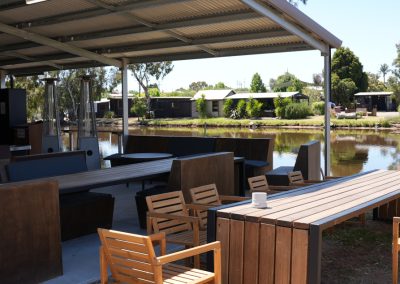Outdoor kitchen facilities at Stawell Hotel Motel Accommodation, featuring a spacious covered area with modern seating arrangements and dining tables. The setup includes patio heaters and a view of the adjacent pond, surrounded by lush greenery and cabins in the background, creating a relaxing social environment