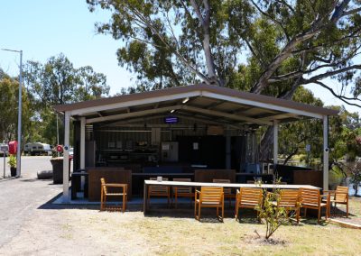Exterior view of the kitchen facilities at Stawell Hotel Motel Accommodation, featuring a spacious covered area with wooden dining tables and chairs. The kitchen setup includes modern amenities and is surrounded by trees and greenery, with a serene pond in the background, creating an inviting space for outdoor dining