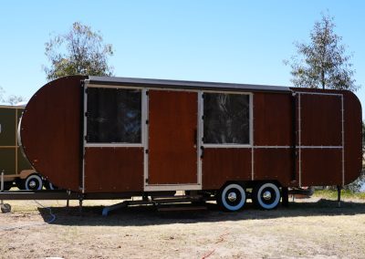 Front view of a stylish brown Hammervan at Stawell Hotel Motel Accommodation, featuring a modern design with rounded edges and multiple entry points. The trailer is set against a backdrop of lush greenery and a serene pond, providing a unique and inviting accommodation option for guests