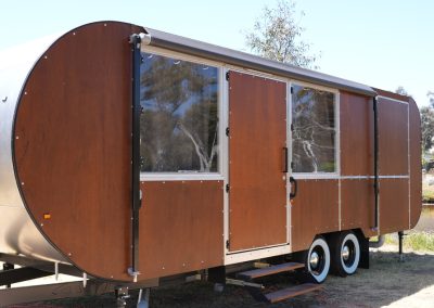 Side view of a stylish brown Hammervan at Stawell Hotel Motel Accommodation, featuring a modern design with rounded edges and multiple entry points. The trailer is set against a backdrop of lush greenery and a serene pond, providing a unique and inviting accommodation option for guests