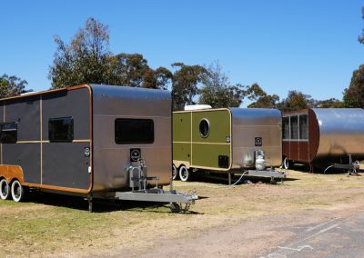 Side view of a row of Hammervans at Stawell Hotel Motel Accommodation, showcasing a mix of modern designs in various colors and finishes. The lineup includes a sleek gray van, a vibrant green van with a circular window, and a unique rounded trailer, all set against a clear blue sky and lush trees in the background