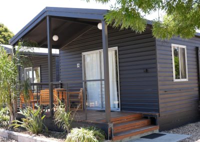 "Front view of Cabin 32 at Stawell Hotel Motel Accommodation, featuring a modern design with a dark exterior and a welcoming porch. The cabin is surrounded by lush greenery, including palm plants, and has outdoor seating for guests to enjoy. Soft lighting and large windows enhance the inviting atmosphere