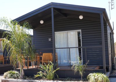 Detailed view of Cabin 32 at Stawell Hotel Motel Accommodation, showcasing its modern design with a dark exterior and a spacious porch. The cabin features outdoor seating, lush greenery including palm plants, and large windows adorned with sheer curtains, enhancing the cozy and inviting atmosphere