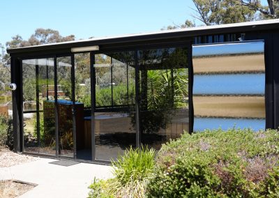 Exterior view of the kitchen facilities at Stawell Hotel Motel Accommodation, featuring large glass windows that provide a bright and open atmosphere. The modern kitchen area includes sleek surfaces and is surrounded by lush greenery, creating a welcoming environment for guests to prepare meals