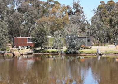 Scenic view of Hammervans at Stawell Hotel Motel Accommodation, set along the water's edge. The image features a variety of unique trailer designs in earthy tones, surrounded by lush greenery and tall trees. The tranquil pond reflects the vans, creating a picturesque and inviting environment for guests