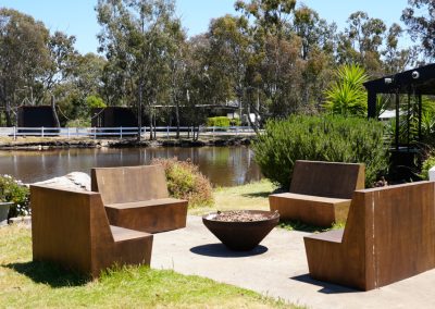 Outdoor seating area at Stawell Hotel Motel Accommodation, featuring modern metal benches arranged around a fire pit. The seating overlooks a tranquil pond, surrounded by lush greenery and trees, creating a cozy and inviting atmosphere for relaxation and social gatherings