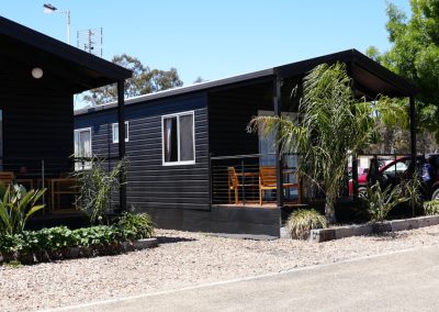 Exterior view of Cabin 32 at Stawell Hotel Motel Accommodation, featuring a sleek dark exterior with a welcoming porch. The cabin is surrounded by lush greenery and palm plants, with outdoor seating visible on the porch. A clear blue sky enhances the inviting atmosphere of this modern accommodation