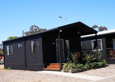 Front view of Cabin 31 at Stawell Hotel Motel Accommodation, featuring a modern dark exterior and a welcoming porch. The cabin has large windows that let in natural light and is surrounded by lush landscaping, including palm plants. A clear blue sky enhances the inviting atmosphere of this cozy accommodation