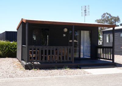 Exterior view of Cabin 30 at Stawell Hotel Motel Accommodation, featuring a stylish dark exterior with a wooden porch. The cabin has large windows and outdoor seating, surrounded by gravel landscaping and greenery. A clear blue sky adds to the inviting atmosphere of this modern accommodation