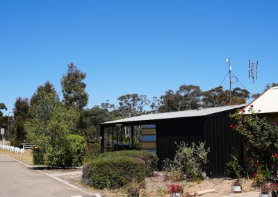 View of the kitchen facilities at Stawell Hotel Motel Accommodation, showcasing a modern building with a mix of dark and light materials. The structure is surrounded by lush greenery and flowers, with a clear blue sky overhead. The setting offers a tranquil ambiance, perfect for outdoor cooking and dining