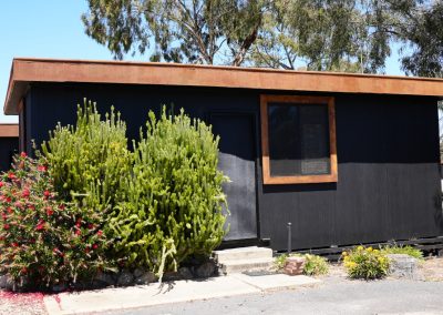 Exterior view of Cabin 28 at Stawell Hotel Motel Accommodation, featuring a modern design with a dark exterior and rustic wooden roof accents. The cabin is surrounded by vibrant greenery, including flowering shrubs and cacti, creating a welcoming atmosphere. The clear blue sky enhances the appeal of this cozy accommodation