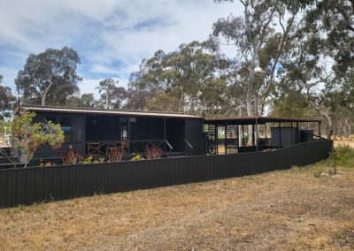 Exterior view of Cabin 88 at Stawell Hotel Motel Accommodation, featuring a modern design with dark walls and a spacious porch. The cabin is surrounded by natural landscaping and tall trees, creating a serene environment. Outdoor seating is visible on the porch, inviting guests to relax and enjoy the peaceful surroundings