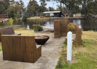 Outdoor seating area at Stawell Hotel Motel Accommodation, featuring modern metal benches arranged around a fire pit. The seating overlooks a tranquil pond, surrounded by lush greenery and trees, creating a cozy atmosphere perfect for relaxation and social gatherings.