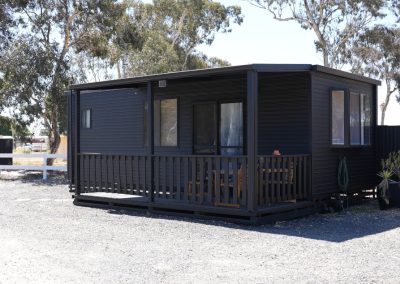 Exterior view of Cabin 91 at Stawell Hotel Motel Accommodation, featuring a modern dark exterior with a welcoming porch. The cabin has large windows that provide natural light and outdoor seating, surrounded by gravel landscaping and lush greenery, creating a cozy and inviting atmosphere
