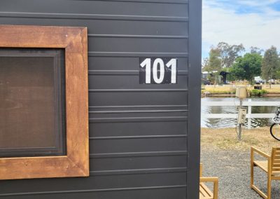 Close-up view of Cabin 101 at Stawell Hotel Motel Accommodation, featuring a sleek dark exterior with a wooden-framed window. The cabin number is prominently displayed next to the window, with a serene view of the pond and outdoor seating area visible in the background, creating a welcoming atmosphere