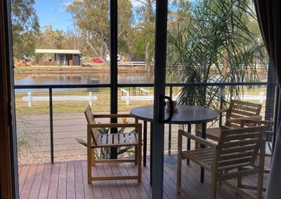 View from inside Cabin 32 at Stawell Hotel Motel Accommodation, showcasing a spacious balcony with outdoor seating. The image captures the inviting wooden deck, a round table, and lush greenery outside, framed by large glass doors that provide a picturesque view of the serene pond and natural surroundings