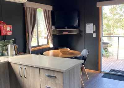 Interior view of Cabin 32 at Stawell Hotel Motel Accommodation, featuring a modern kitchenette with a compact countertop and cabinetry. The space includes a dining area with a round table and chairs, along with a mounted TV. Large windows allow natural light to fill the room, providing a cozy and inviting atmosphere