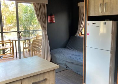 Interior view of Cabin 32 at Stawell Hotel Motel Accommodation, showcasing a cozy living space with a comfortable sofa and kitchenette. The room features a modern design with a refrigerator and storage cabinets, along with large sliding glass doors that lead to a balcony with outdoor seating, allowing natural light to brighten the area