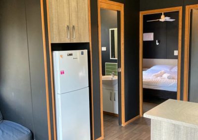 Interior view of Cabin 32 at Stawell Hotel Motel Accommodation, showing the kitchenette area with a refrigerator and cabinetry. The space features a modern design, with a comfortable seating area visible in the corner, and doorways leading to a bathroom and bedroom. Natural light enhances the cozy atmosphere of this well-appointed cabin