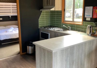 Interior view of the kitchen area in Cabin 32 at Stawell Hotel Motel Accommodation, showcasing modern cabinetry and a gas stove with an exhaust hood. The design features green tiles and a countertop that seamlessly connects to the dining area. A doorway leads to the bedroom, enhancing the cabin's inviting and functional layout