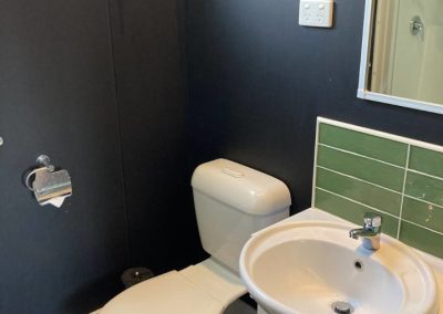 Interior view of the bathroom in Cabin 32 at Stawell Hotel Motel Accommodation, featuring a modern toilet and a sink with a circular basin. The bathroom includes stylish green tiles, a large mirror, and a window for natural light, creating a clean and functional space for guests
