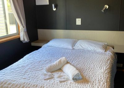 Interior view of the bedroom in Cabin 32 at Stawell Hotel Motel Accommodation, featuring a comfortable double bed with crisp white linens and neatly arranged towels. The room includes modern lighting fixtures and a ceiling fan, with large windows allowing natural light to brighten the space, creating a cozy and inviting atmosphere