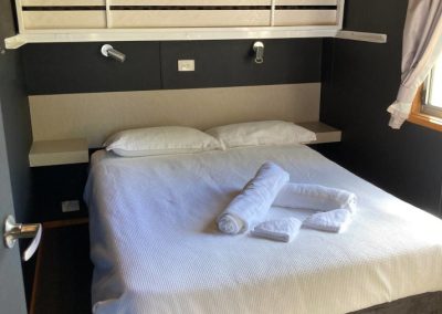 Close-up view of the bedroom in Cabin 32 at Stawell Hotel Motel Accommodation, featuring a neatly made bed with white linens and arranged towels. The room includes a modern headboard design, wall-mounted reading lights, and a cozy ambiance created by the dark wall colors and natural light coming through the window