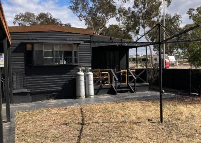 Exterior view of Cabin 88 at Stawell Hotel Motel Accommodation, showcasing a modern black cabin with a wooden deck and gas tanks in the foreground. The cabin features large windows and is surrounded by natural vegetation, set against a backdrop of partly cloudy skies, creating a serene outdoor atmosphere