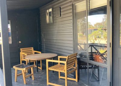 Porch view of Cabin 88 at Stawell Hotel Motel Accommodation, featuring a wooden table surrounded by chairs. The porch is covered and showcases a sleek black exterior, with a door leading to the cabin's interior. The space is designed for relaxation, overlooking a serene outdoor environment