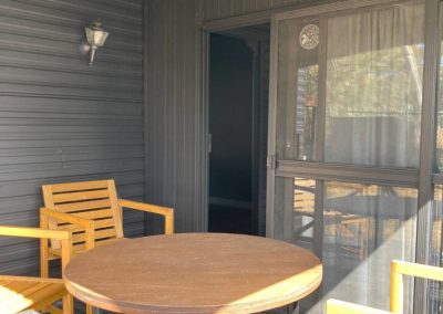 Cozy porch area of Cabin 88 at Stawell Hotel Motel Accommodation, featuring a round wooden table and two wooden chairs. The cabin's exterior is painted in a sleek black finish, with a door leading into the cabin's interior. A wall-mounted light fixture enhances the inviting atmosphere