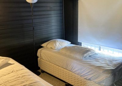 Interior view of Cabin 88, showcasing a cozy bedroom with two beds: a double bed on the left with a thick comforter and a single bed on the right. A floor lamp stands beside the single bed, and a window covered with a white blind allows natural light to enter the room at Stawell Hotel Motel Accommodation