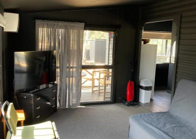 Interior view of Cabin 88, featuring a cozy living area with a gray fold-out couch and a green folding chair. A large flat-screen TV is positioned against the wall, complemented by a black dresser. Natural light filters in through the curtains, revealing a glimpse of the outdoor deck with seating. A small kitchen area is visible in the background, along with a vacuum cleaner and trash bin at Stawell Hotel Motel Accommodation