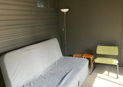 Cozy interior of Cabin 88 featuring a gray fold-out couch with a simple design, paired with a green folding chair and two small wooden side tables. A standing lamp provides light in the corner, creating a comfortable and inviting atmosphere. The walls are painted in a dark tone, enhancing the warm ambiance at Stawell Hotel Motel Accommodation