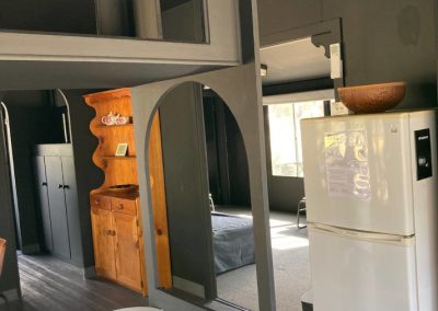 Interior view of Cabin 88 showcasing a modern layout. The space features a minimalist design with dark walls and wooden accents. A white countertop is visible in the foreground, while a stylish wooden shelving unit is seen against the wall. In the background, a cozy sleeping area is visible with a gray fold-out couch and a refrigerator, contributing to a functional yet inviting atmosphere at Stawell Hotel Motel Accommodation