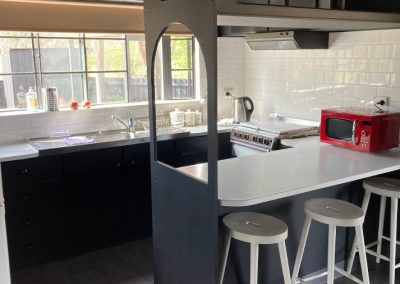 Modern kitchen interior of Cabin 88 featuring sleek design elements. The kitchen includes a white countertop with bar stools, a stainless steel sink, and a red microwave. Above the counter, open shelving displays various kitchen items. Natural light floods the space through large windows, highlighting the contemporary white tile backsplash and black cabinetry that create a stylish and functional cooking area at Stawell Hotel Motel Accommodation