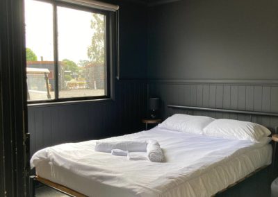 Interior view of Cabin 116's bedroom featuring a cozy, neatly made bed with white linens. Two towels are carefully arranged on the bed. The room is brightly lit with a fluorescent light fixture on the ceiling and has a large window that allows natural light to enter, enhancing the warm and inviting atmosphere. A small bedside table with a lamp sits next to the bed, contributing to the room's comfort at Stawell Hotel Motel Accommodation