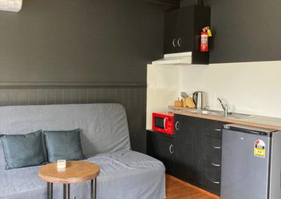 Interior view of Cabin 116's kitchenette and living area, showcasing a cozy gray sofa with decorative pillows. A small wooden side table sits beside the sofa, adding functionality to the space. The kitchenette is equipped with a modern sink, a microwave oven, and a refrigerator, featuring dark cabinetry for a sleek appearance. A bright fluorescent light fixture illuminates the area, enhancing the inviting ambiance of the cabin at Stawell Hotel Motel Accommodation