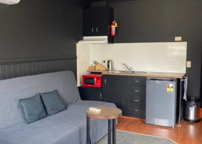 Interior view of Cabin 116's living space, featuring a compact kitchen area equipped with a sink, kettle, microwave, and refrigerator. A gray sofa with two decorative pillows is situated in front of a small round table. The walls are painted dark, and a light fixture illuminates the room, creating a cozy atmosphere at Stawell Hotel Motel Accommodation