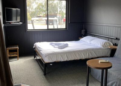 Cozy interior of Cabin 116 showcasing a neatly made bed with fresh linens and rolled towels. A small round table sits beside the bed, while a gray sofa is positioned nearby. The room features dark walls, a large window allowing natural light to flood in, and a wall-mounted television, creating a welcoming atmosphere at Stawell Hotel Motel Accommodation