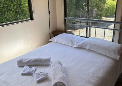 Inviting interior of Cabin 57 featuring a neatly made double bed with white linens and neatly rolled towels. Natural light streams in through large windows, highlighting the cozy atmosphere. A lamp stands beside the bed, and lush greenery is visible outside, enhancing the tranquil setting at Stawell Hotel Motel Accommodation
